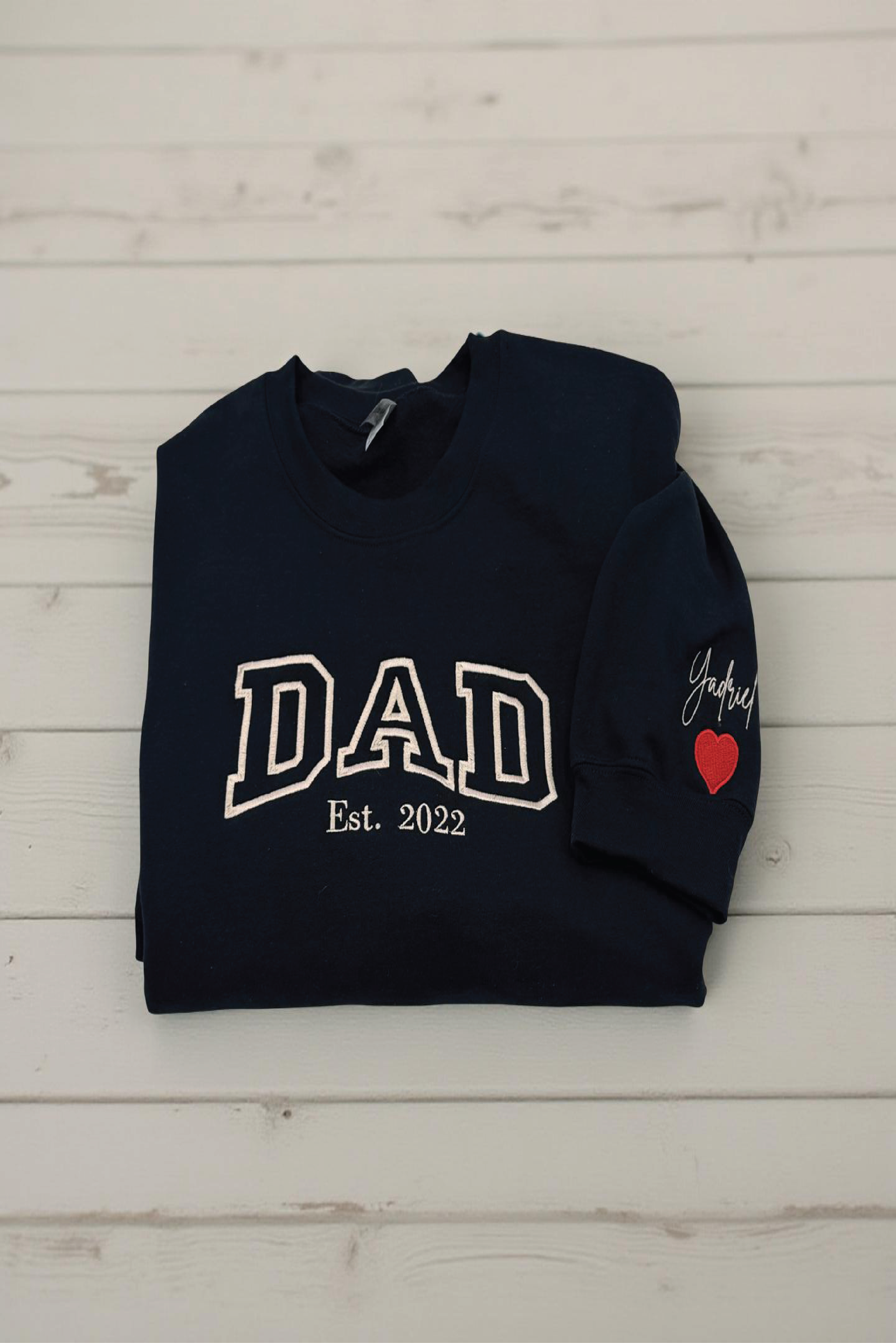 Navy blue sweatshirt with 'DAD Est. 2022' text and a heart on a light wooden background

Dad custom embroidered sweatshirt with kids’ names on sleeve – black pullover

Personalized dad sweatshirt with child name embroidered on sleeve – gray crewneck